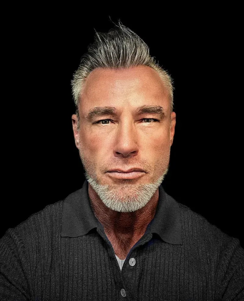 Portrait of a man with gray spiked hair and a salt-and-pepper beard in a dark ribbed polo.